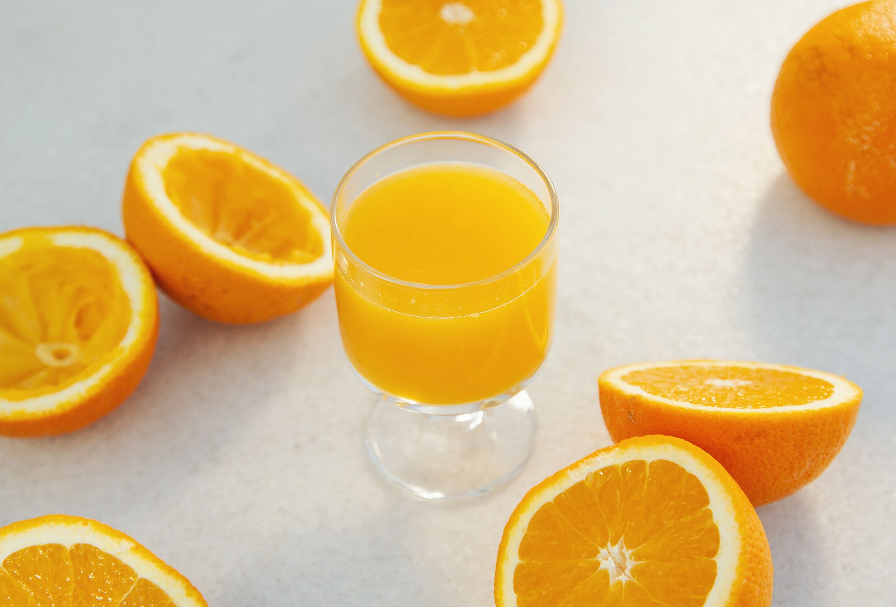 A glass of orange juice surrounded by whole and halved oranges on a light-colored surface.