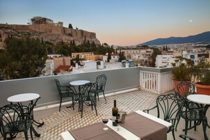 Μια βεράντα με καθίσματα σε εξωτερικό χώρο, ένα μπουκάλι κρασί και θέα σε ένα ιστορικό κτίριο.