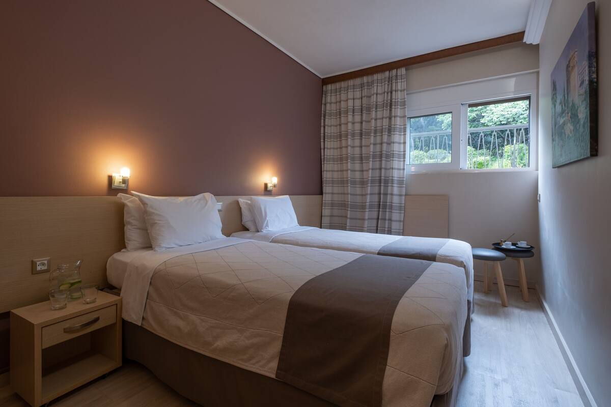 Habitación de hotel con dos camas, una mesita de noche, una pequeña mesa con lámpara y una ventana con cortinas.