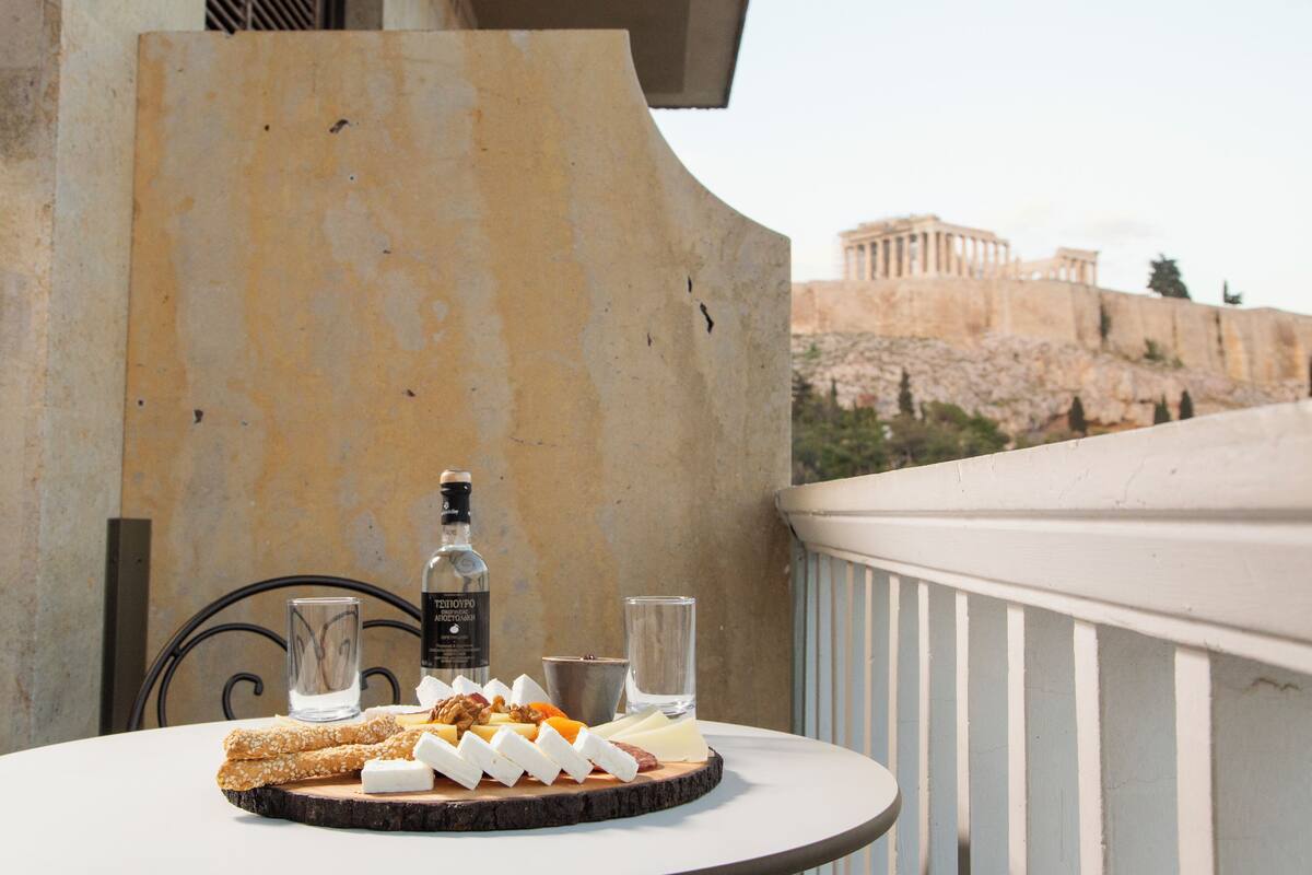 Una mesa con una tabla de quesos, frutos secos y una botella de licor en un balcón con vistas a ruinas antiguas.