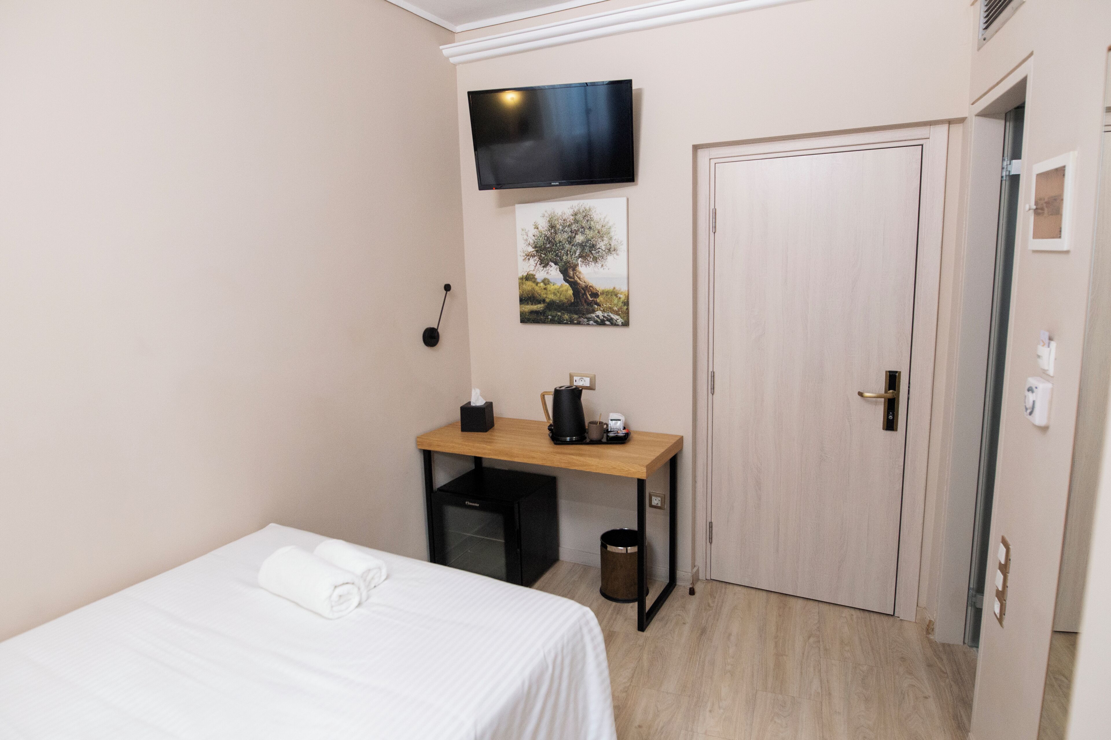 Een hotelkamer met een bed, een bureau met koffiezetapparaat, een prullenbak, een aan de muur bevestigde televisie en een deur.