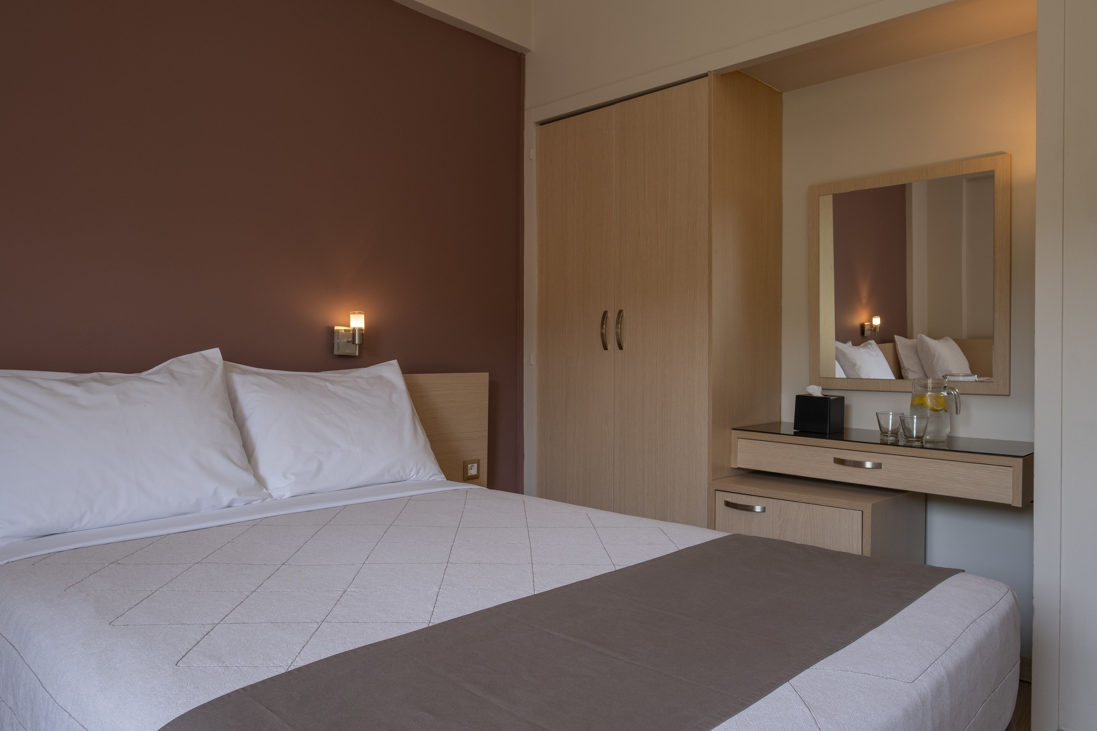Quarto de hotel com uma cama, um guarda-roupa, uma mesa de cabeceira com abajur e um espelho.