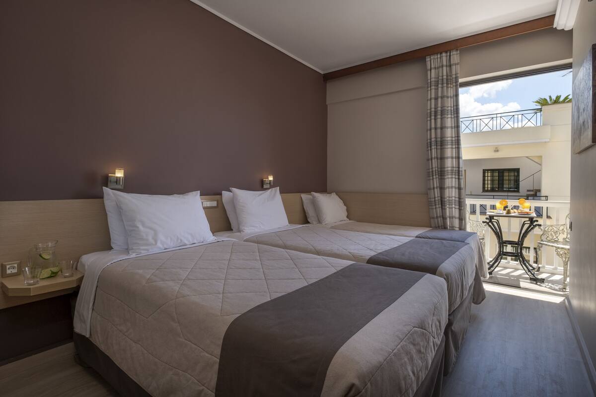 Habitación de hotel con dos camas, una mesita con lámpara y un balcón con vistas.