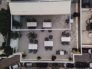 An aerial view of a rooftop patio with tables and chairs arranged in a grid pattern, surrounded by potted plants and a parking area with cars.