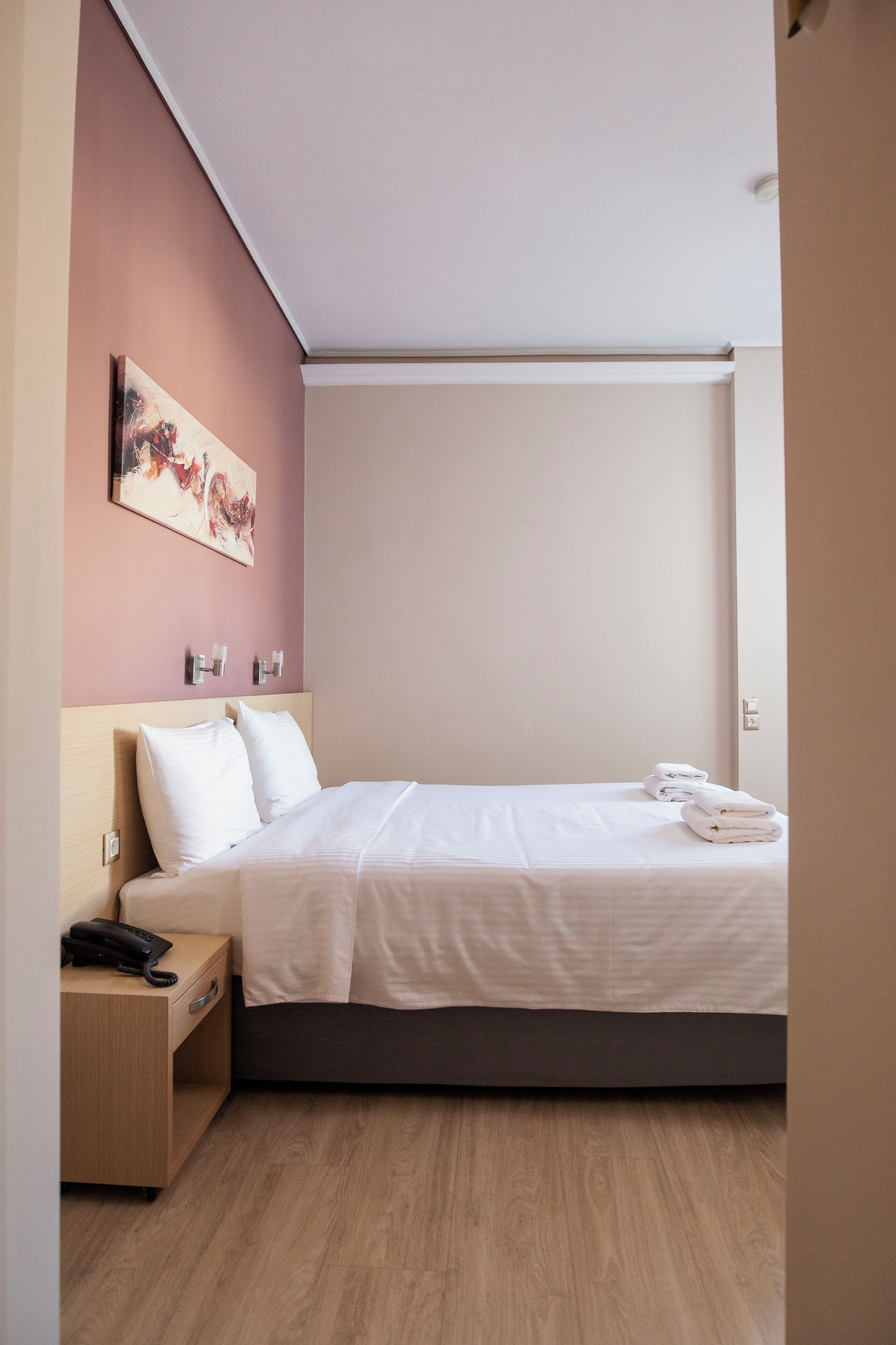 A neatly made bed with white linens, a wooden nightstand with a phone, and a painting on the wall.