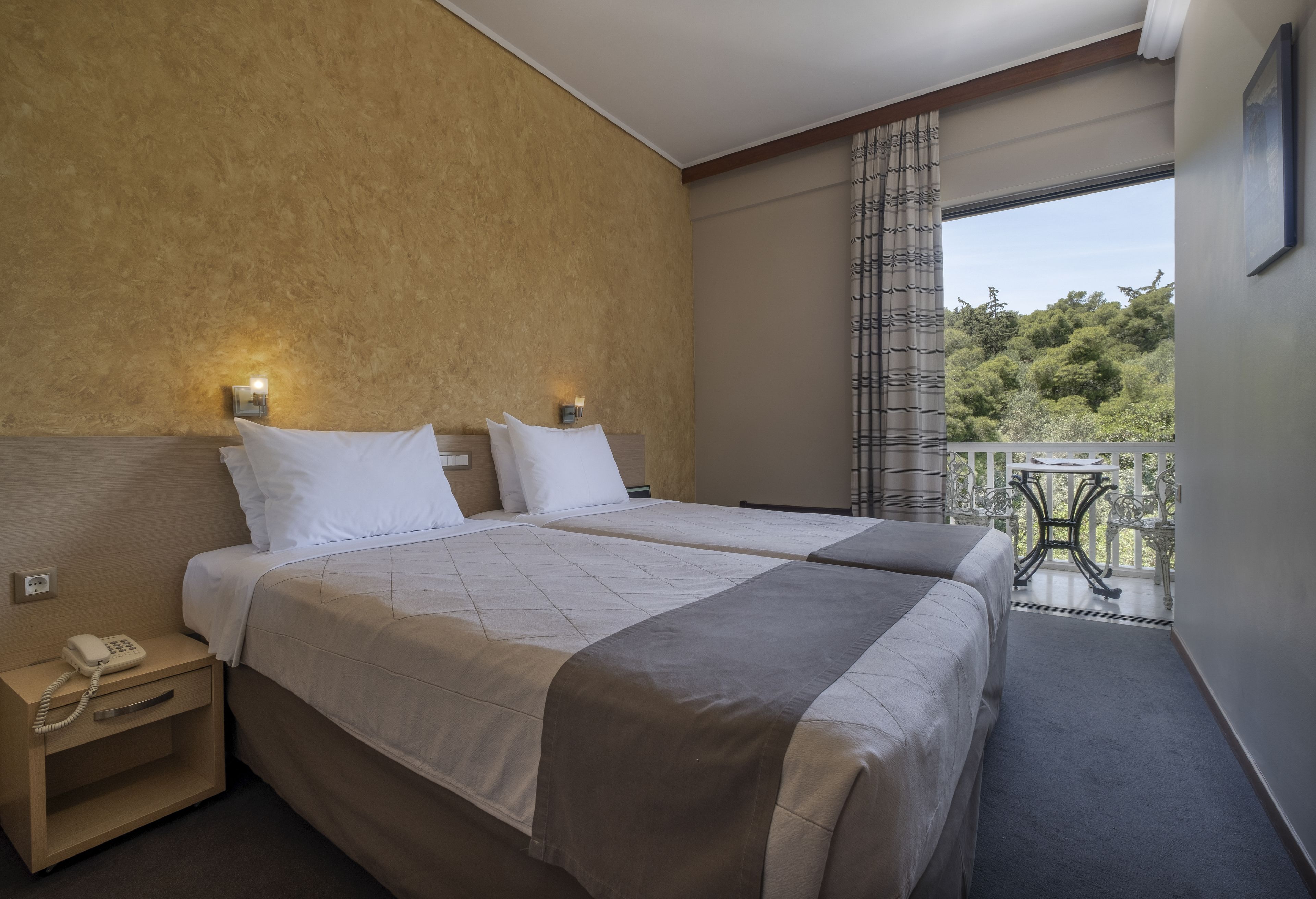 A hotel room with two beds, a nightstand with a phone, a balcony with a table and chairs, and a view of trees.