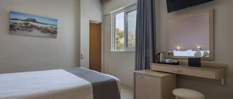 A hotel room with a bed, a desk with a mirror, a TV mounted above the desk, and a window with curtains.