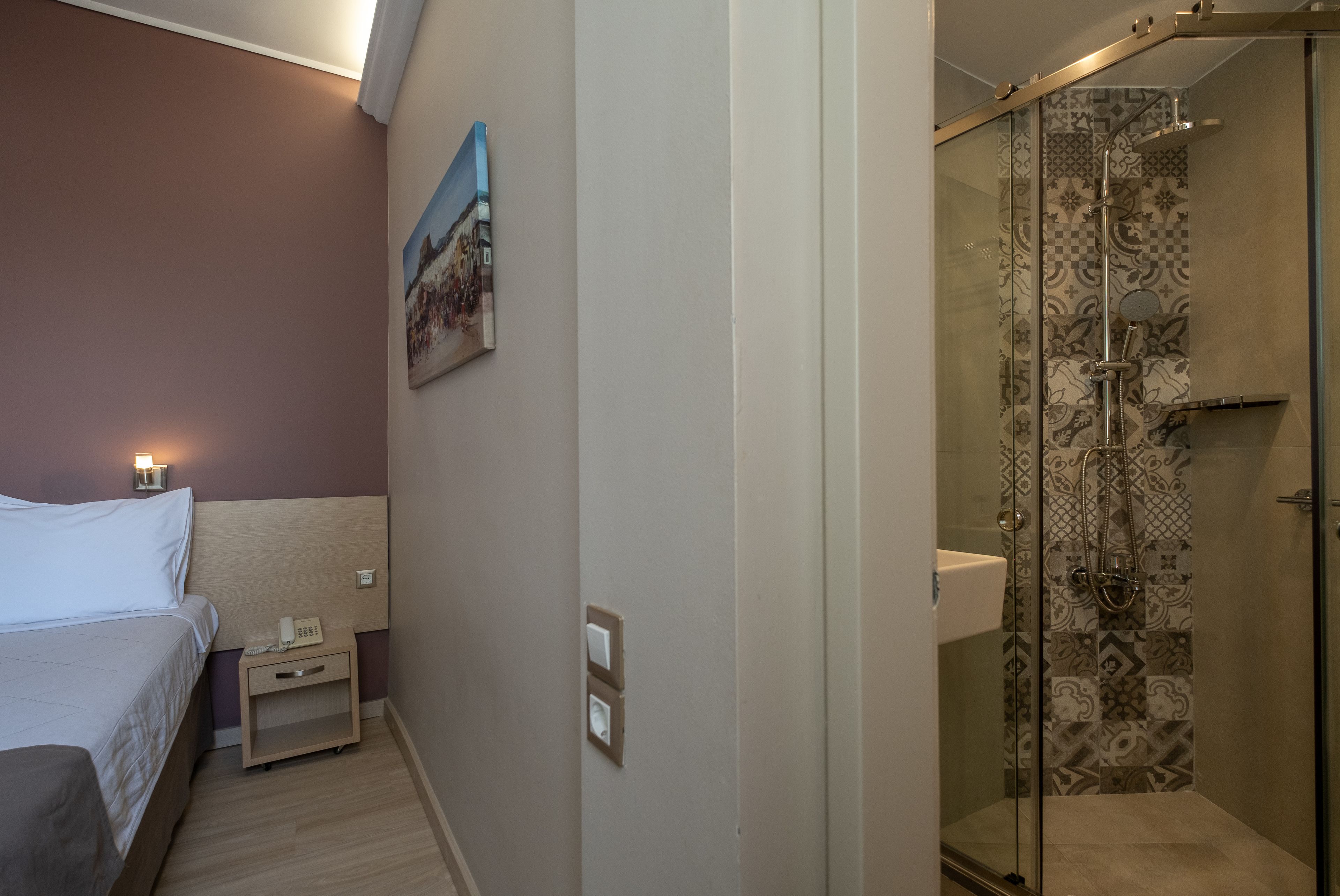 A hotel room with a bed, bedside table with a phone, a painting on the wall, and a shower area with patterned tiles.