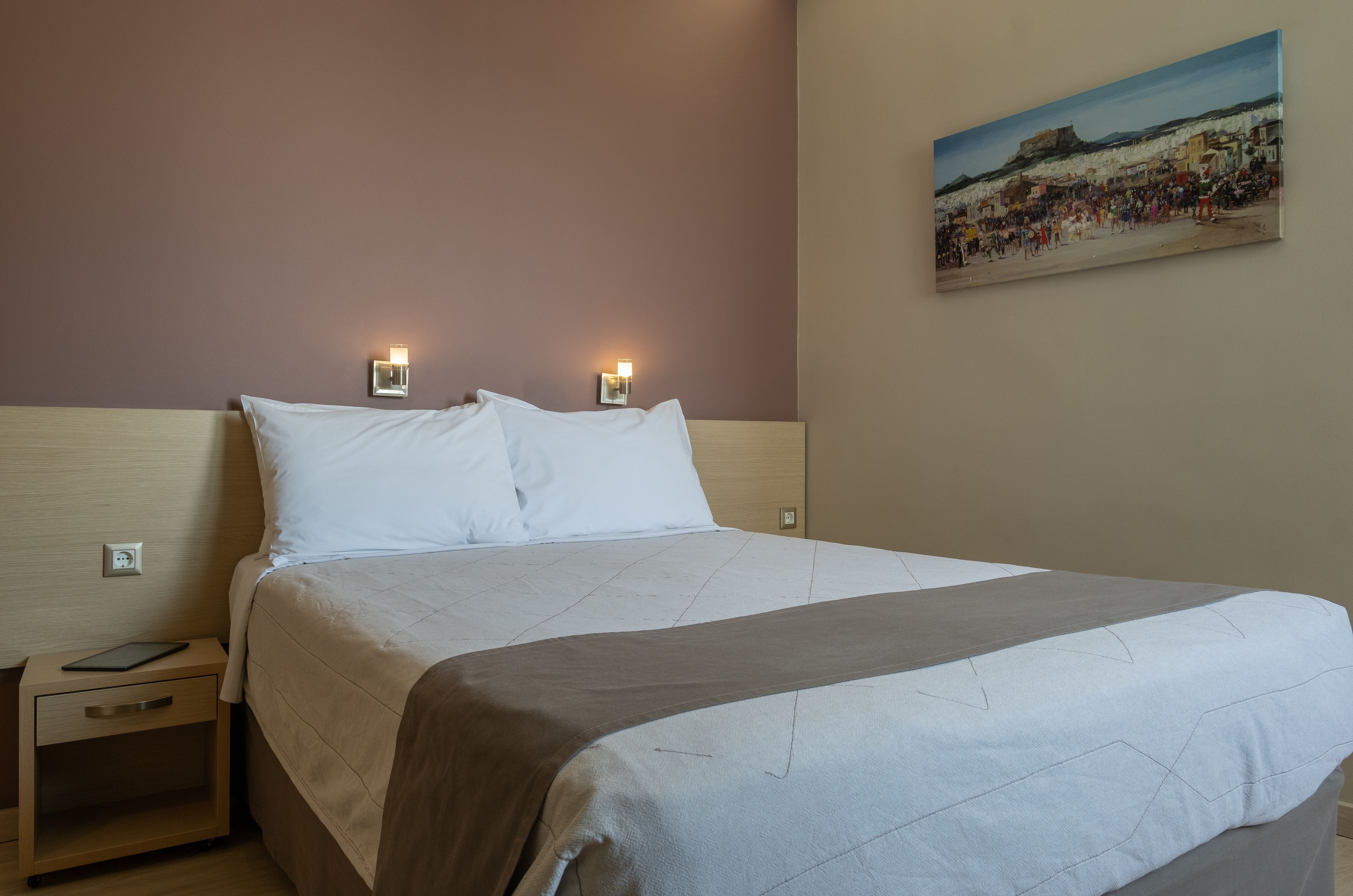 Ein ordentlich bezogenes Bett mit heller Bettdecke und Kissen, ein Nachttisch mit einem Tablet und ein Bild einer Strandlandschaft an der Wand.