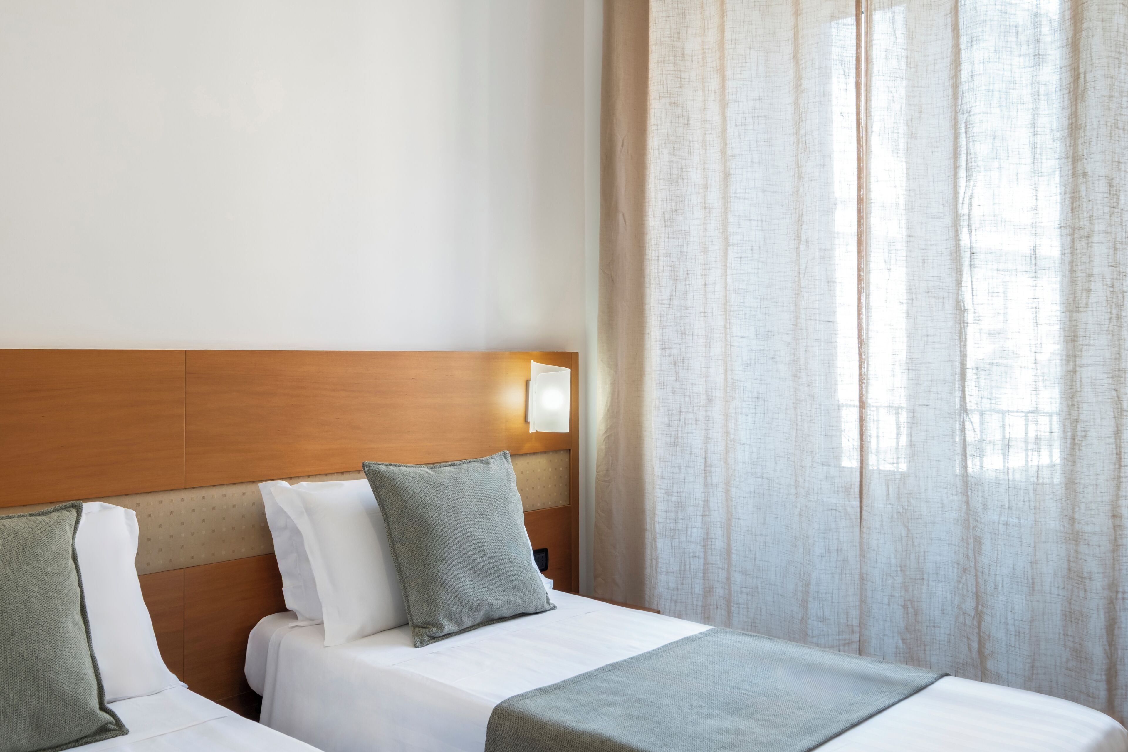 A hotel room with a wooden headboard, two beds, a nightstand, a lamp, and sheer curtains.