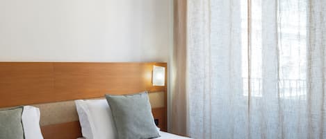 A hotel room with a wooden headboard, two beds, a nightstand, a lamp, and sheer curtains.