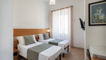 Habitación de hotel con dos camas individuales, cabecero de madera, ventana con cortinas translúcidas, una obra de arte enmarcada en la pared y un televisor de pantalla plana.