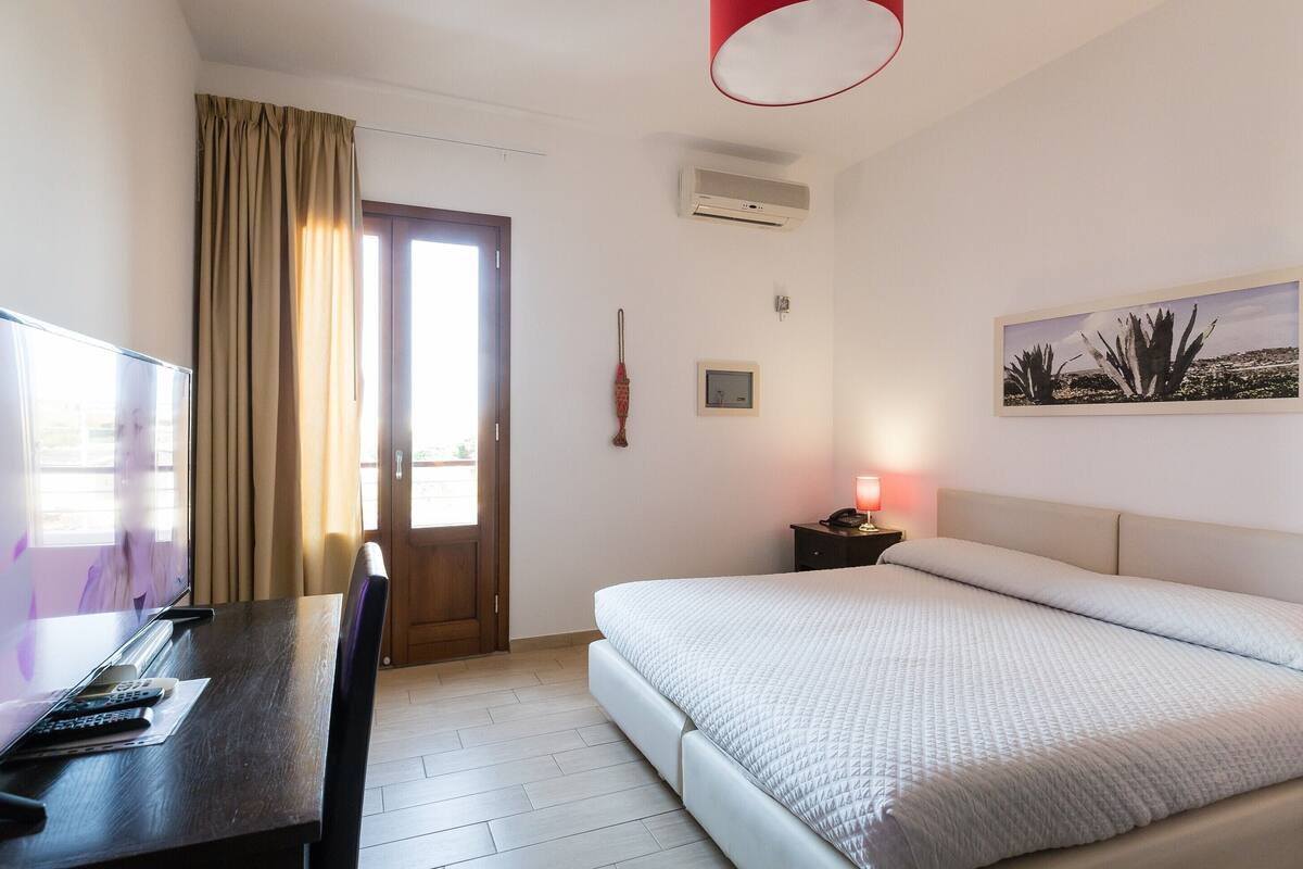 A hotel room with a bed, a desk with a TV, a door, and a wall-mounted air conditioner.