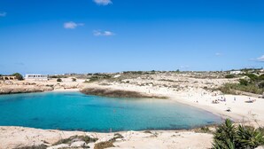 A coastal landscape with a beach, clear blue water, and a white building in the background. - Hotel Ristorante Cavalluccio Marino (Lampedusa)