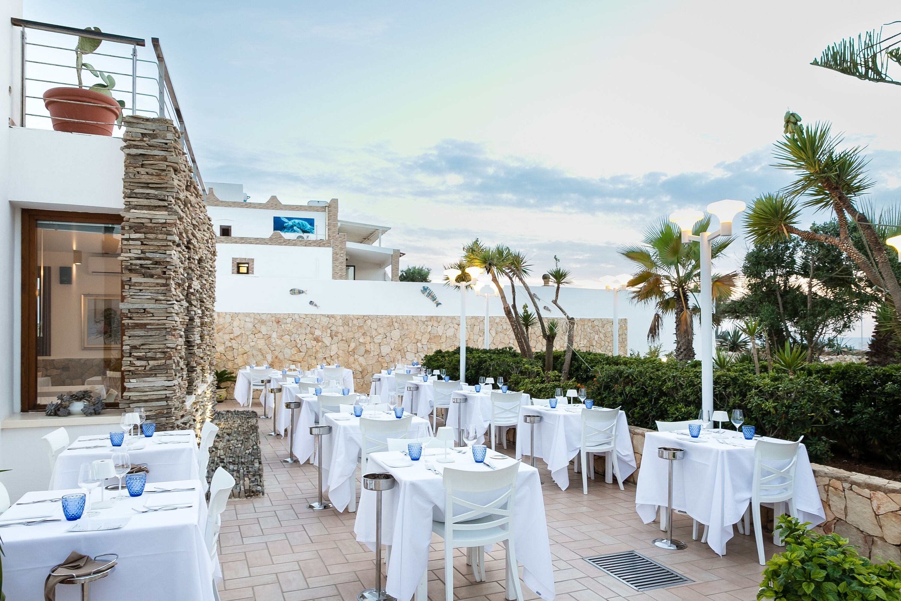 An outdoor dining area with white tables and chairs, stone pillars, and a view of a building with a balcony.