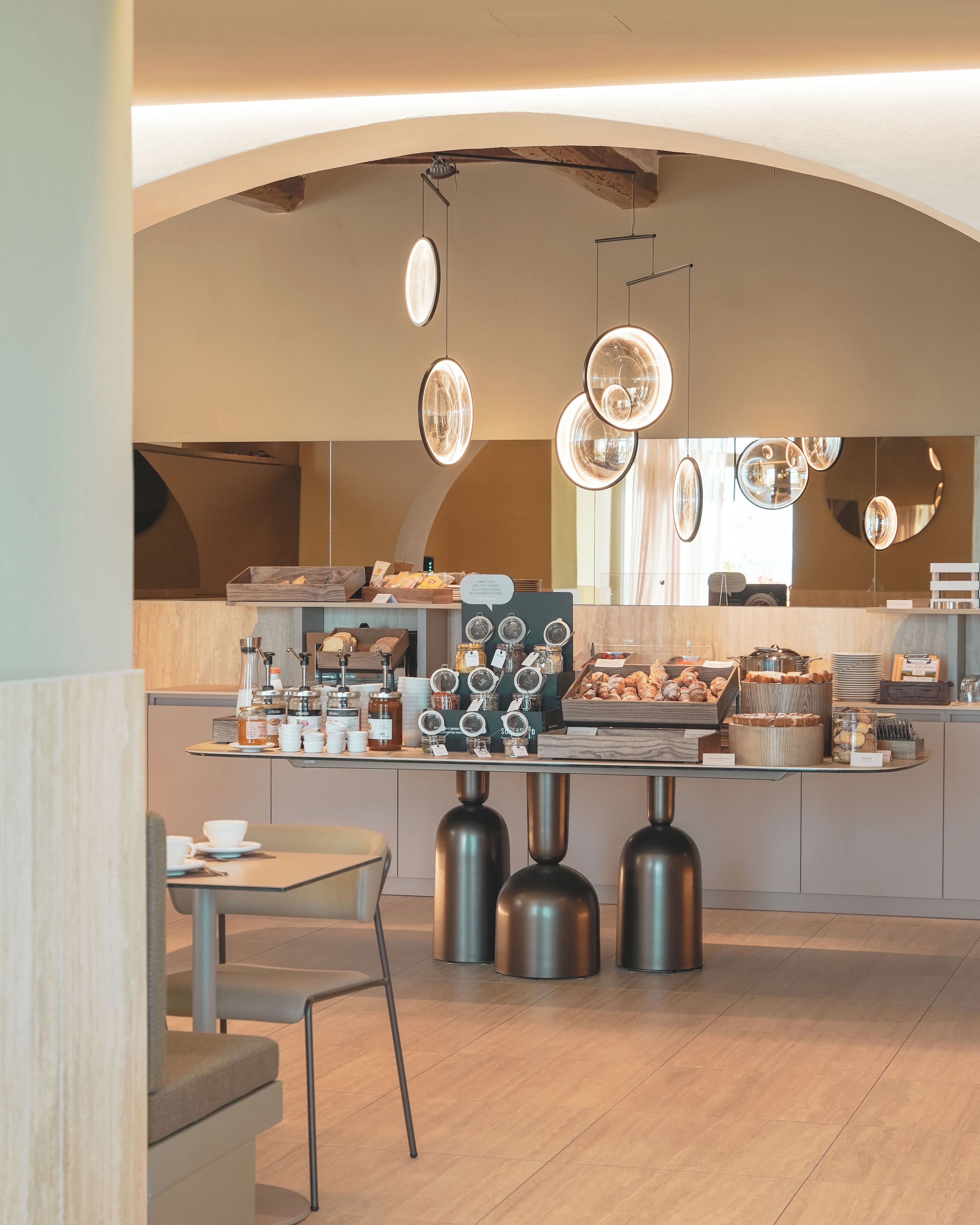 A modern cafe with a central counter displaying pastries and beverages, a seating area with a table and chairs, and unique circular ceiling lights.
