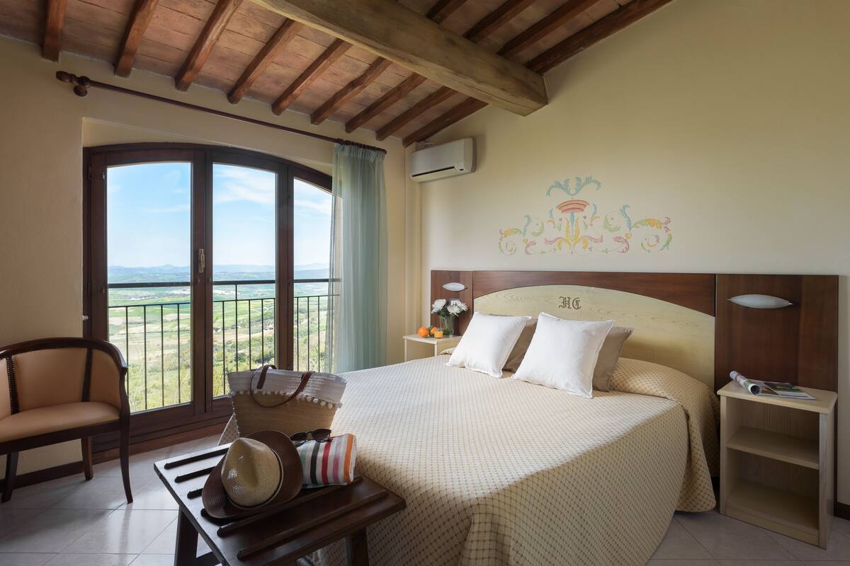 A hotel room with a large bed, a chair, a table with a hat and sunglasses, a balcony with a view, and a wall with a floral mural.