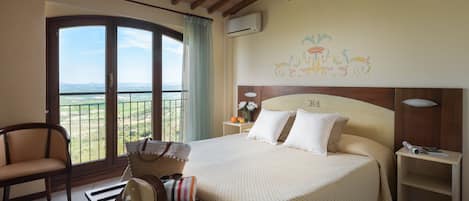 A hotel room with a large bed, a chair, a table with a hat and sunglasses, a balcony with a view, and a wall with a floral mural.