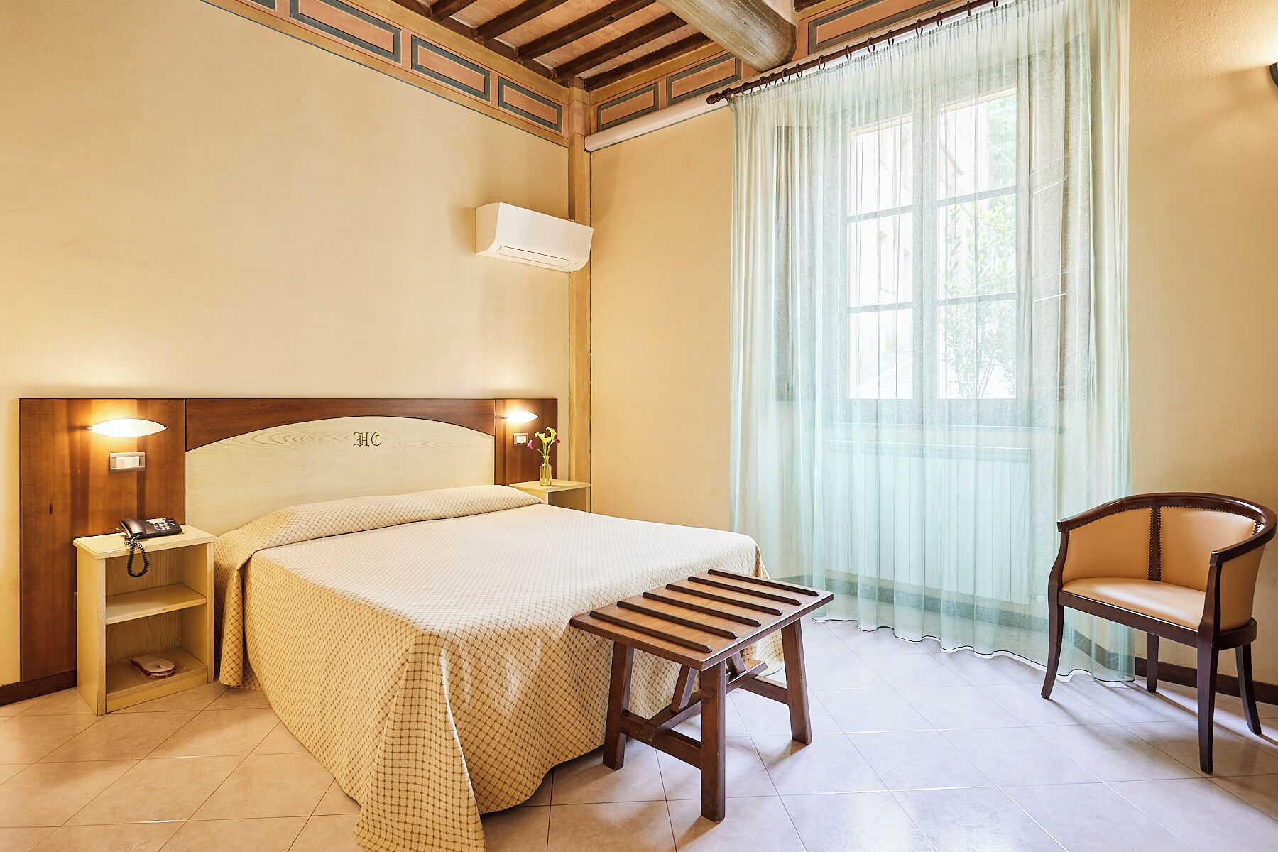 A hotel room with a bed, bedside lamps, a nightstand, a wooden bench, a chair, and a window with sheer curtains.
