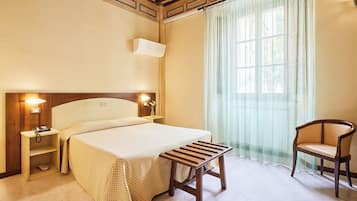 Ein Hotelzimmer mit einem Bett, Nachttischlampen, einem Nachttisch, einer Holzbank, einem Sessel und einem Fenster mit durchscheinenden Vorhängen.