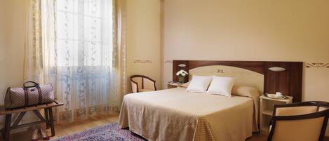 A hotel room with a large bed, a wooden nightstand, a chair, a small table with a vase and a cup, and a patterned rug.