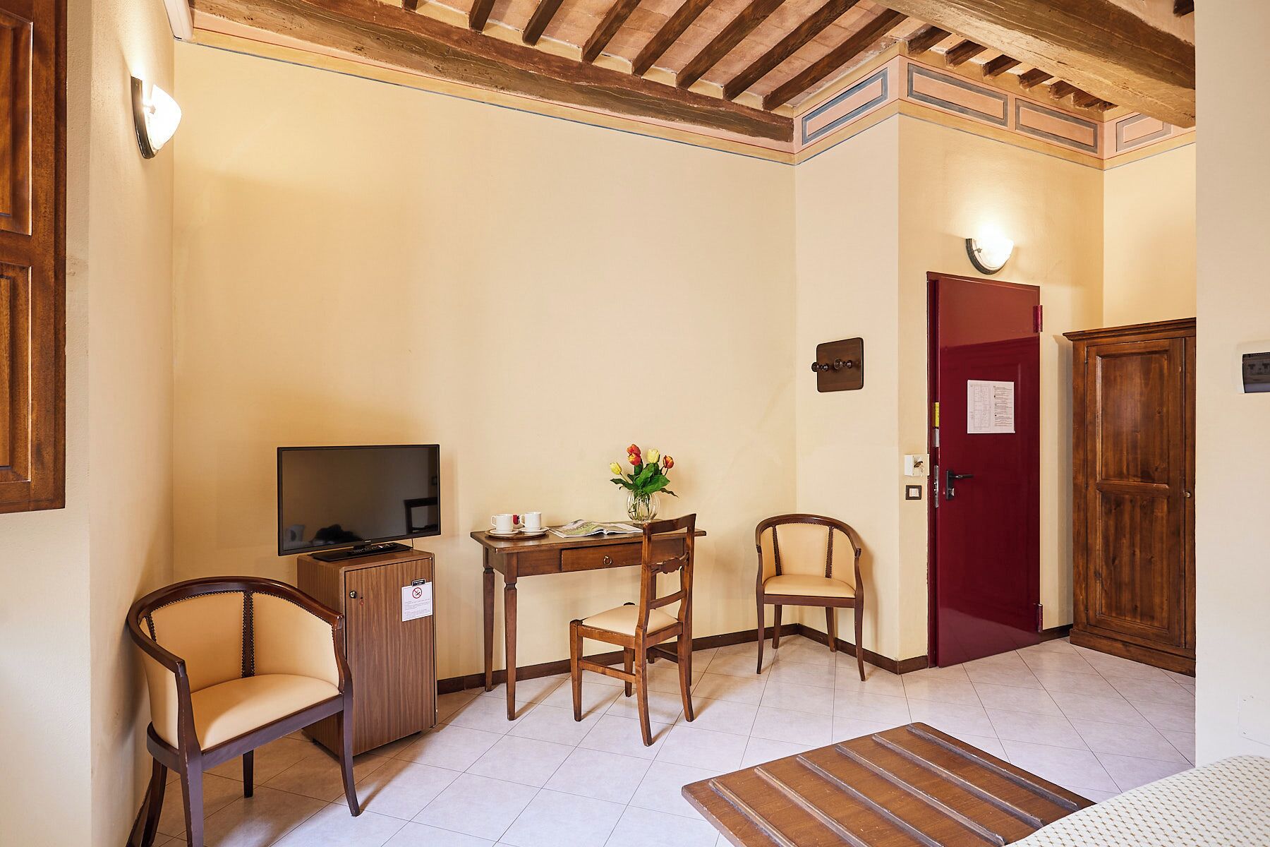 Una habitación pequeña y acogedora con televisión, un escritorio con un florero, dos sillas y una puerta roja.