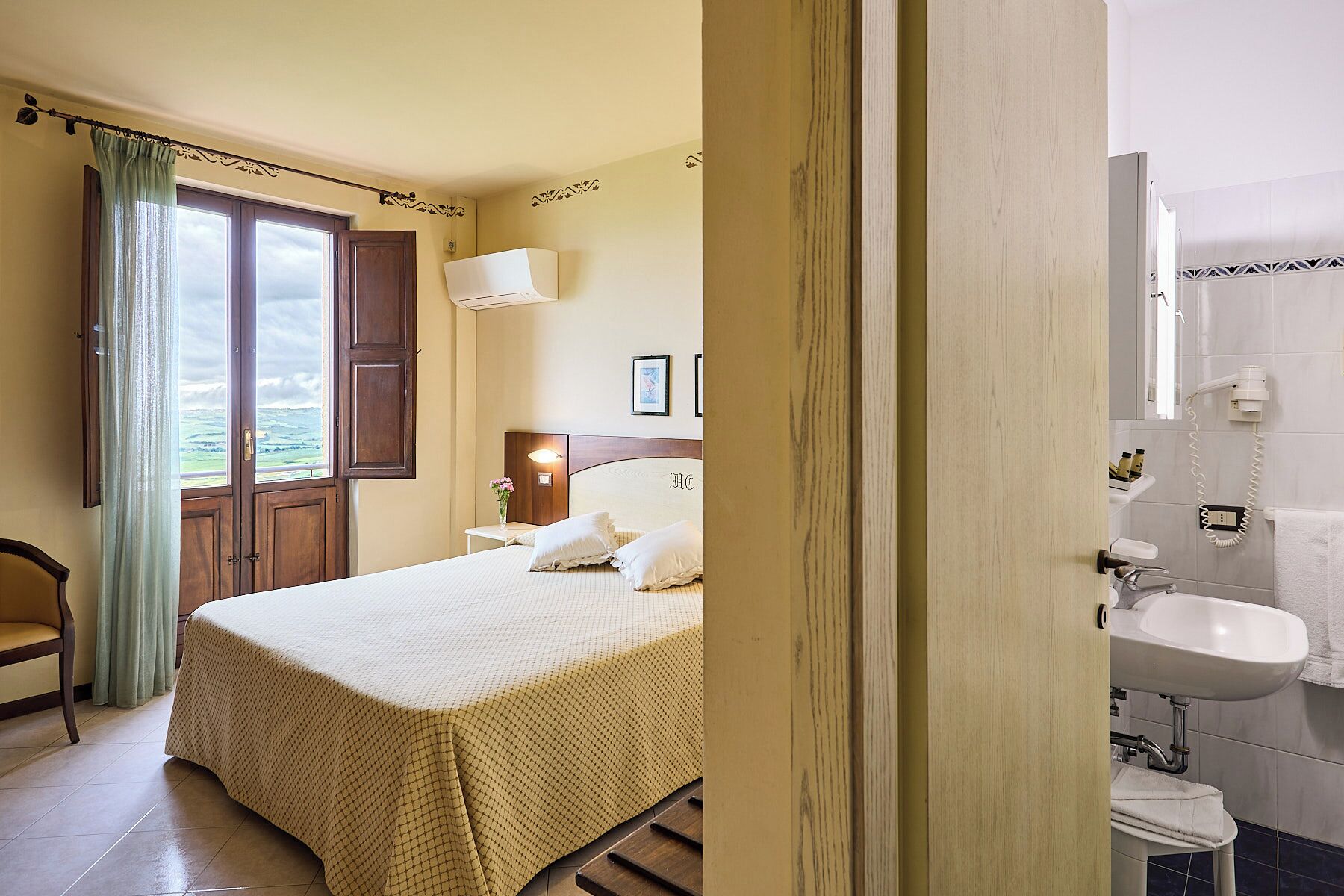 Ein Hotelzimmer mit Bett, Nachttisch, einem Fenster mit Vorhängen, einer Wandlampe und Meerblick.
