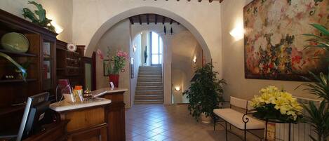Un vestíbulo amplio con suelo de baldosas, un mostrador de recepción de madera, una escalera y un cuadro grande con motivos florales en la pared.