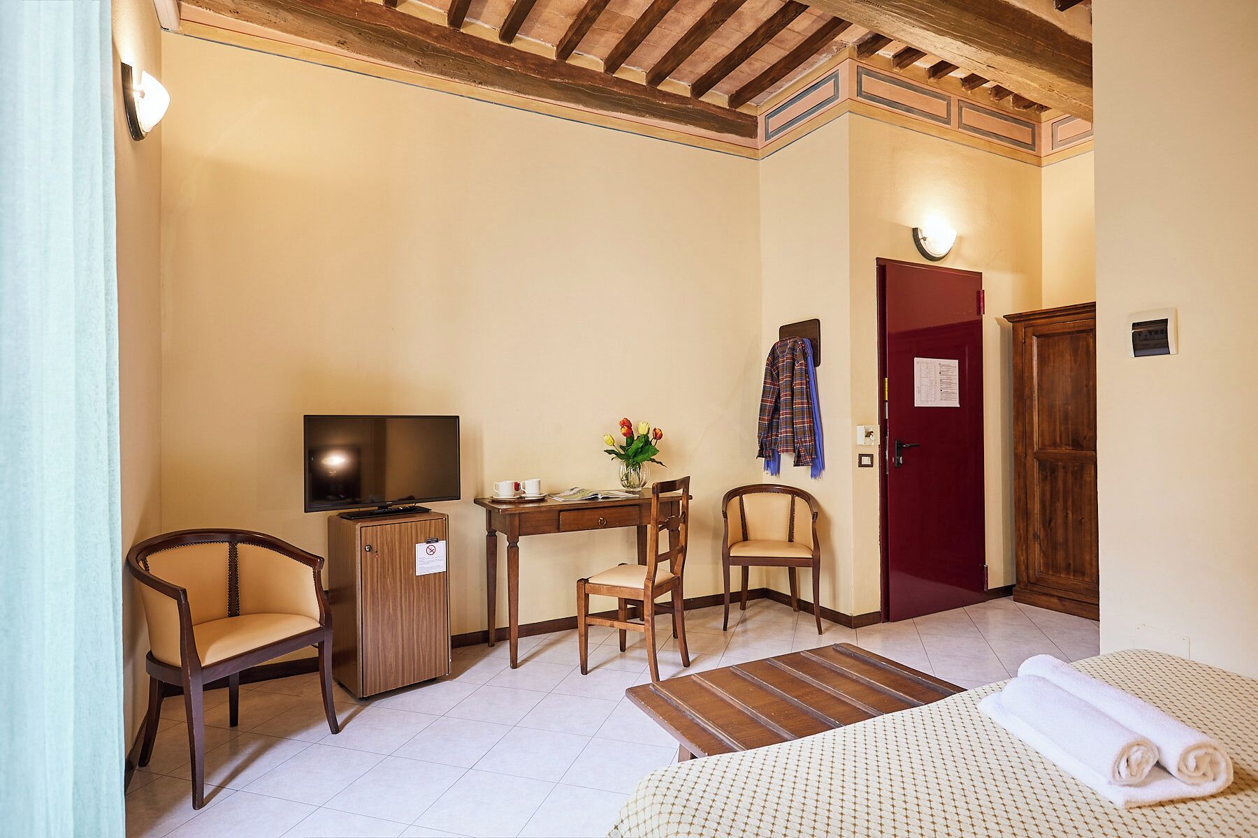 A hotel room with a bed, a desk with a television, two chairs, a small table with a vase of flowers, and a red door.