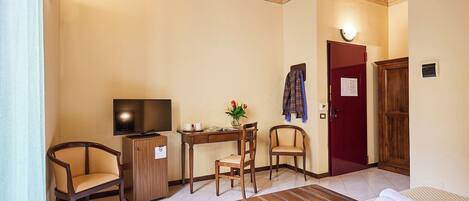 A hotel room with a bed, a desk with a television, two chairs, a small table with a vase of flowers, and a red door.