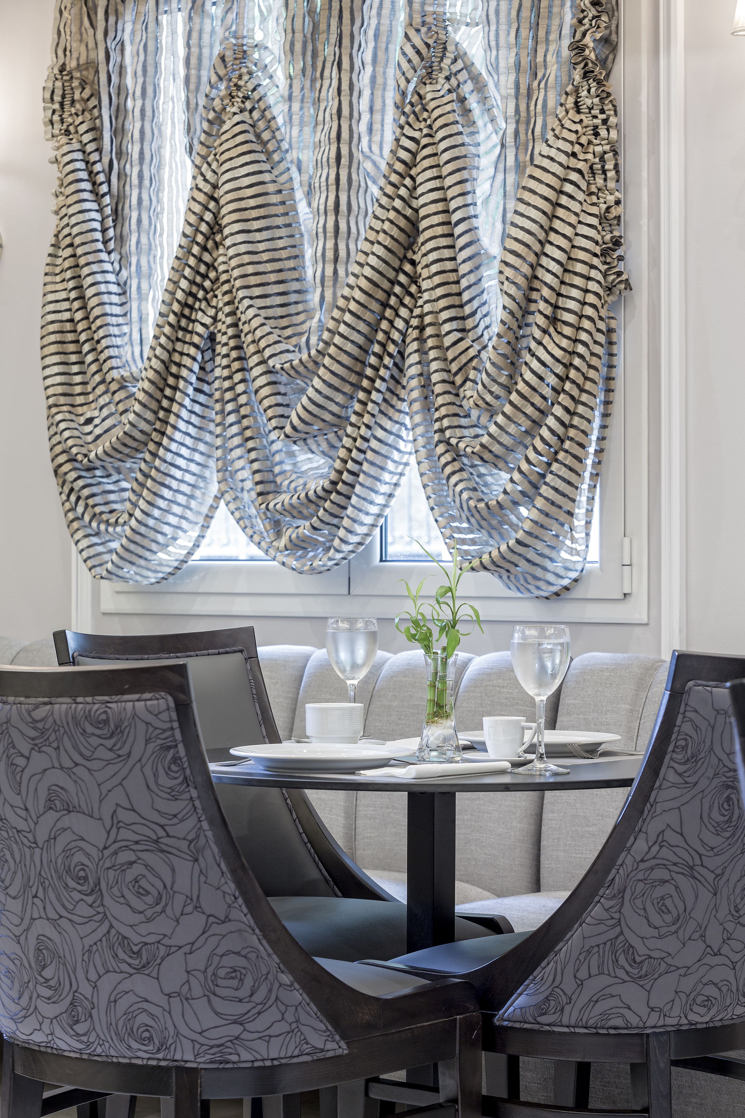 A dining area with a round table, grey upholstered chairs, and a window with striped curtains.