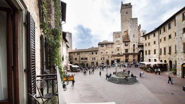 A historic square with a central fountain, surrounded by stone buildings with arched windows and a prominent tower. - La Cisterna (San Gimignano)