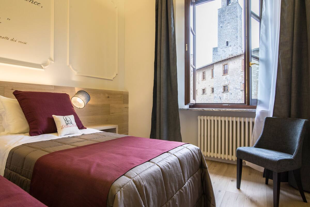 A hotel room with a bed, a chair, a window with a view of a building, and a radiator.