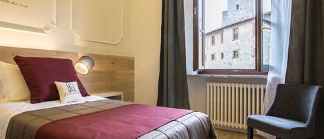 Quarto de hotel com cama, cadeira, janela e vista para um edifício, além de radiador.