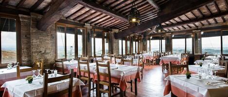 A restaurant with wooden beams, stone walls, and large windows offering a view of the landscape. Tables are set with white tablecloths and red napkins.
