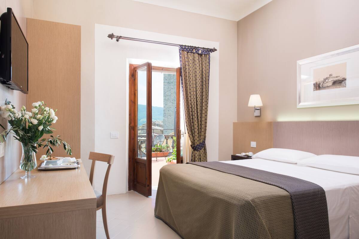 A hotel room with a bed, a desk with a vase of flowers, a chair, a bedside table, a lamp, and a view of the outdoors through a door.