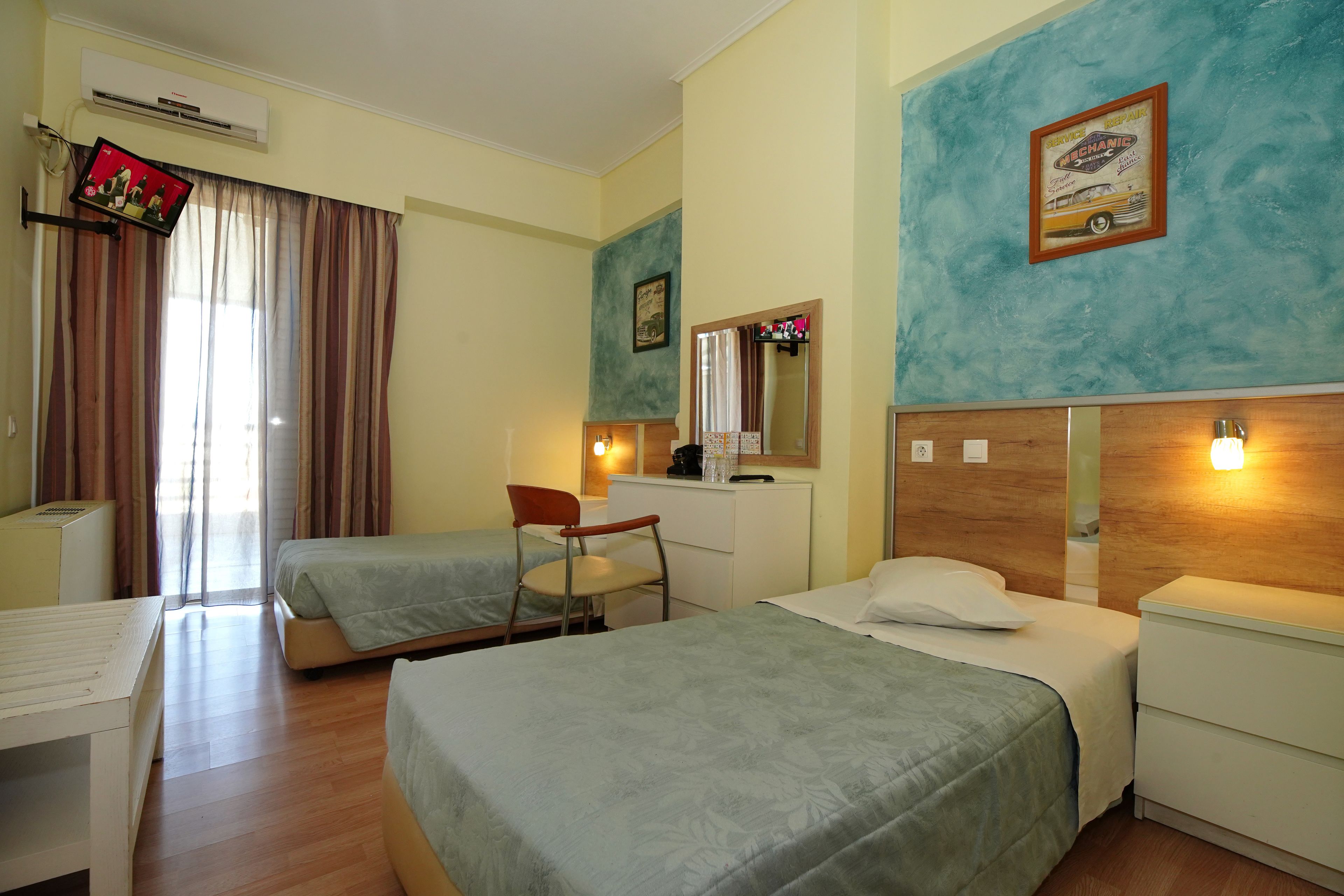 A hotel room with two single beds, a desk, a chair, a small table, a mirror, and a TV mounted on the wall.