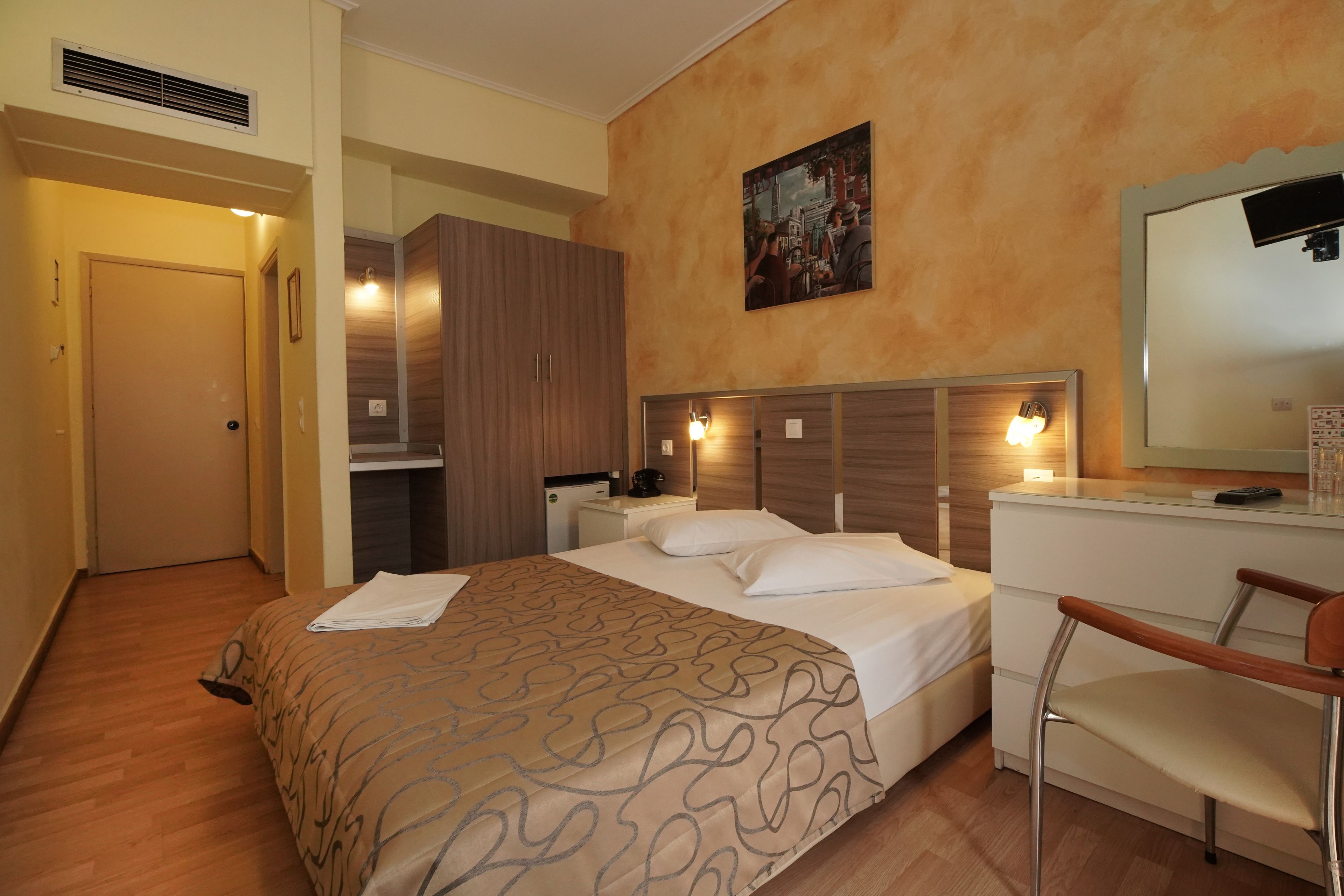 A hotel room with a bed, a desk, a chair, a wardrobe, and a painting on the wall.