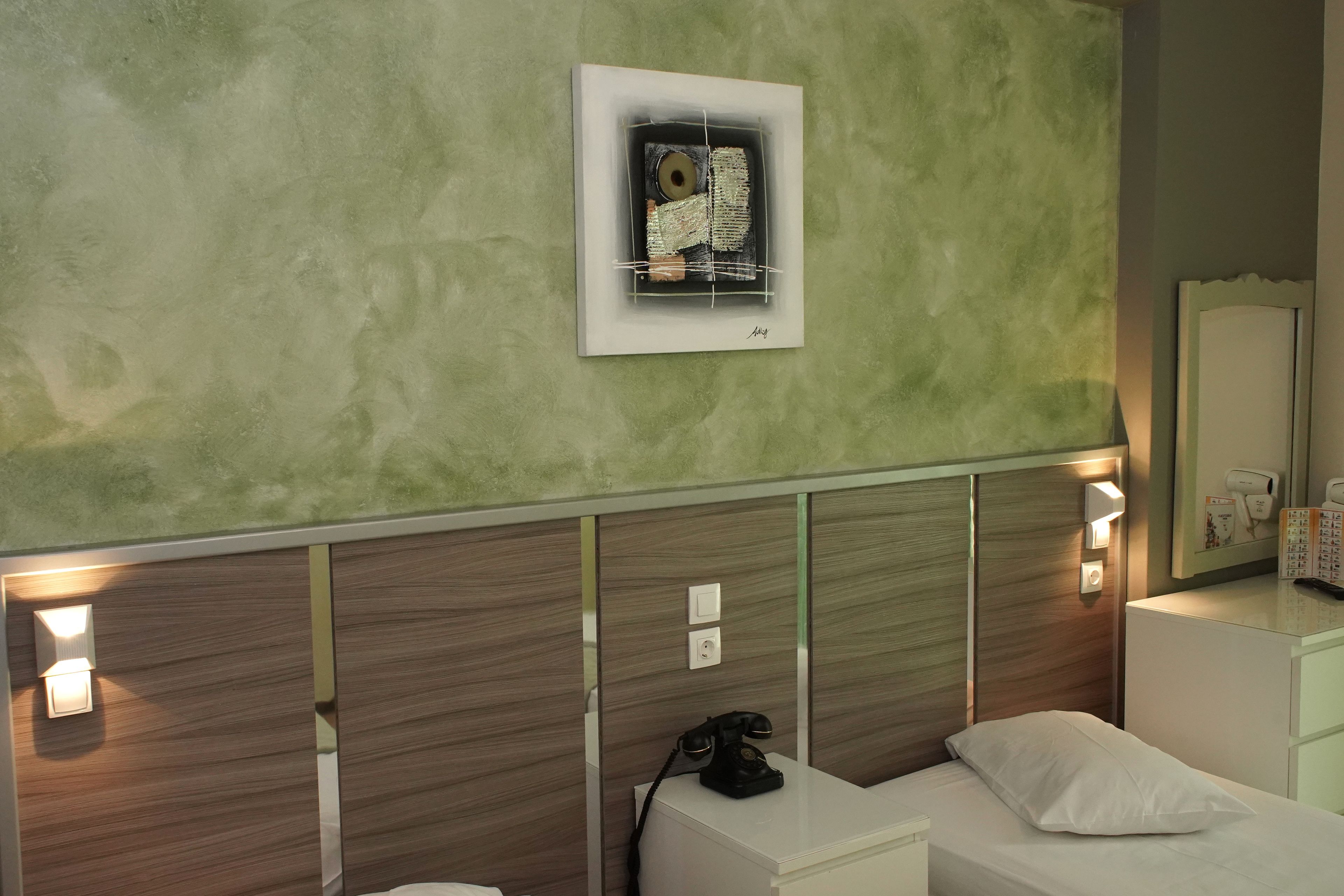 Habitación de hotel con pared verde texturizada, una obra de arte enmarcada, lámparas de noche, un teléfono y una cama blanca.