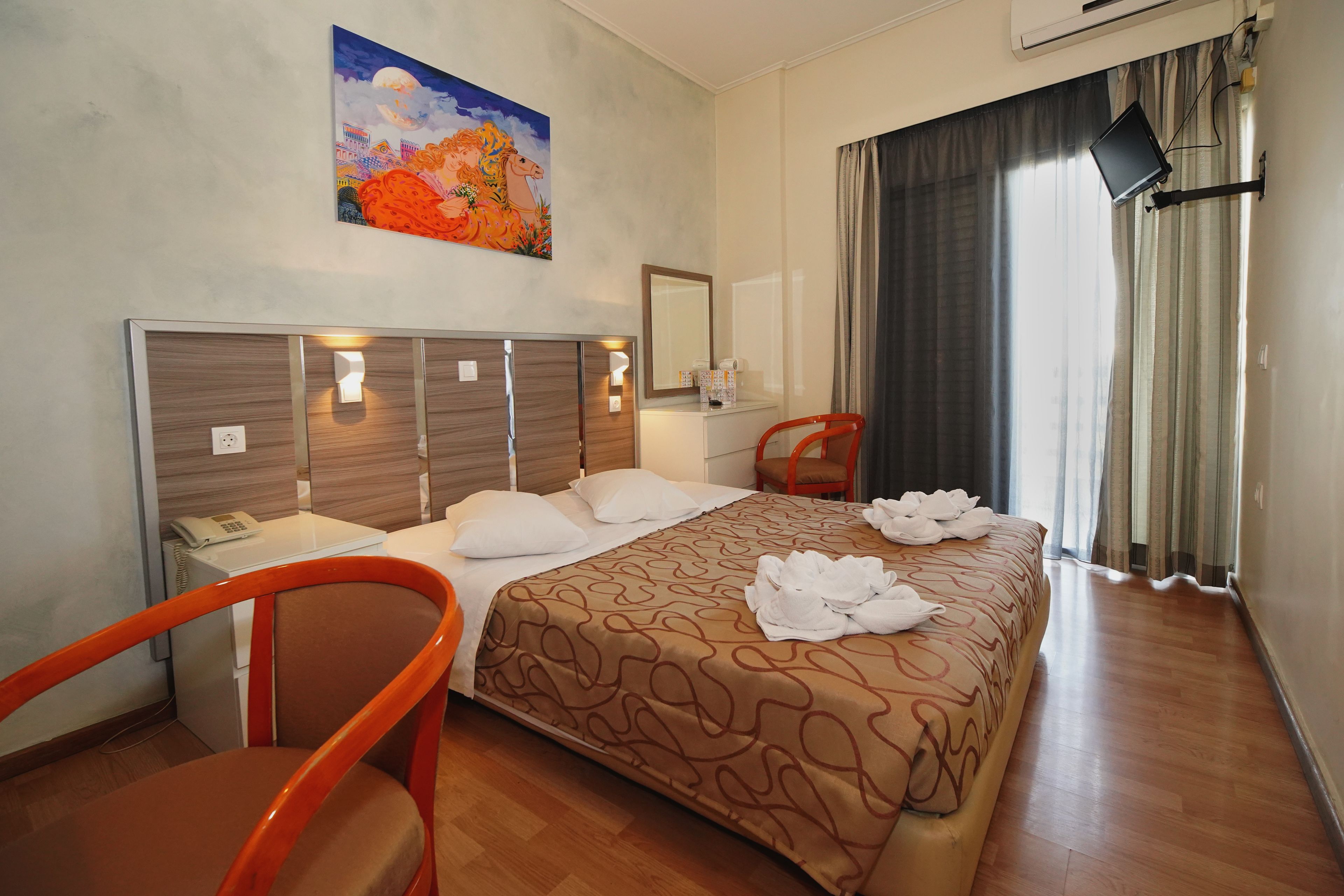 A hotel room with a bed, a desk, a chair, a painting on the wall, and a window with curtains.