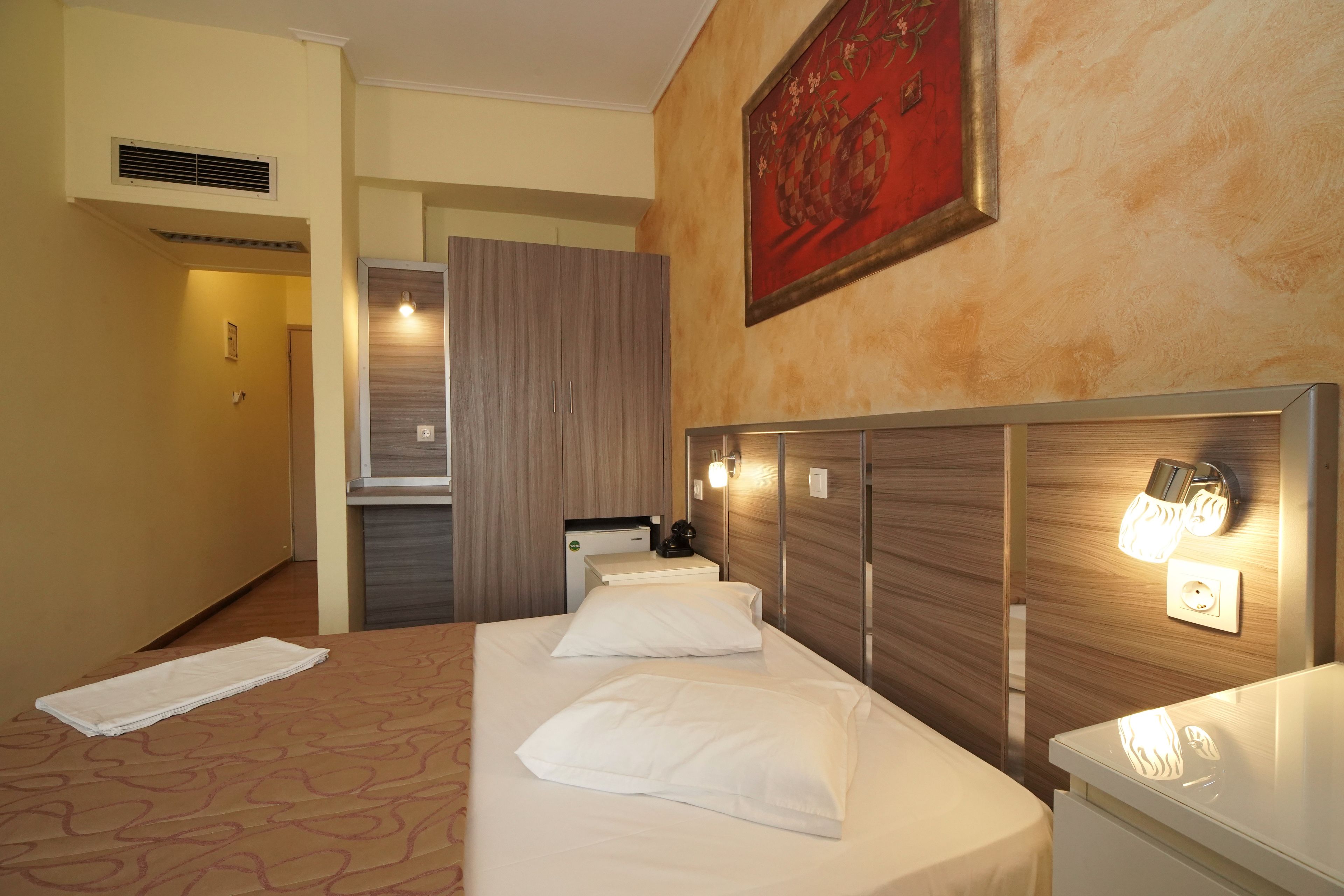 A hotel room with a bed, bedside tables, a nightstand, a wall-mounted lamp, a painting, and a vent.