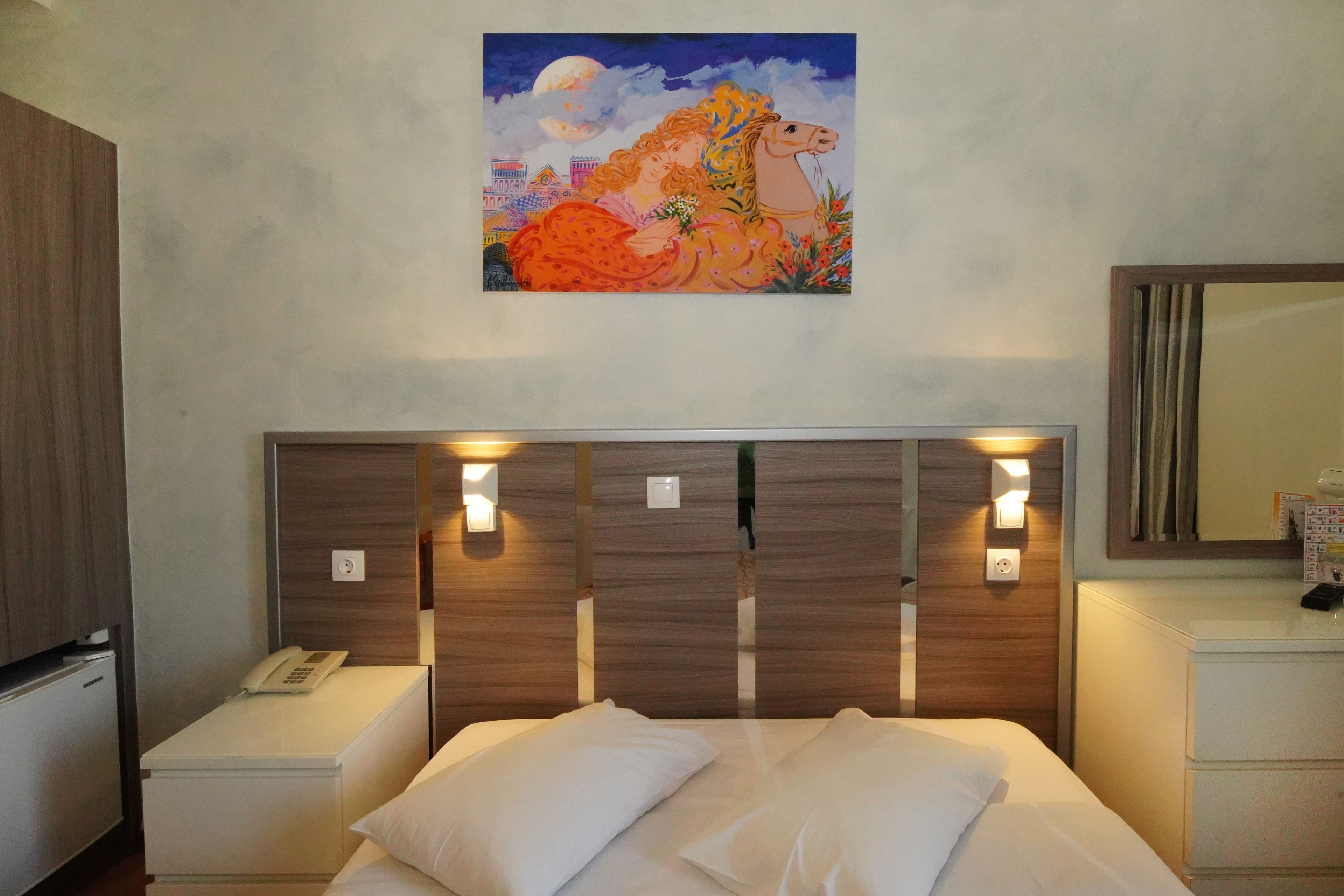A hotel room with a wooden headboard, bedside lamps, a phone, and a painting of a woman with a horse.