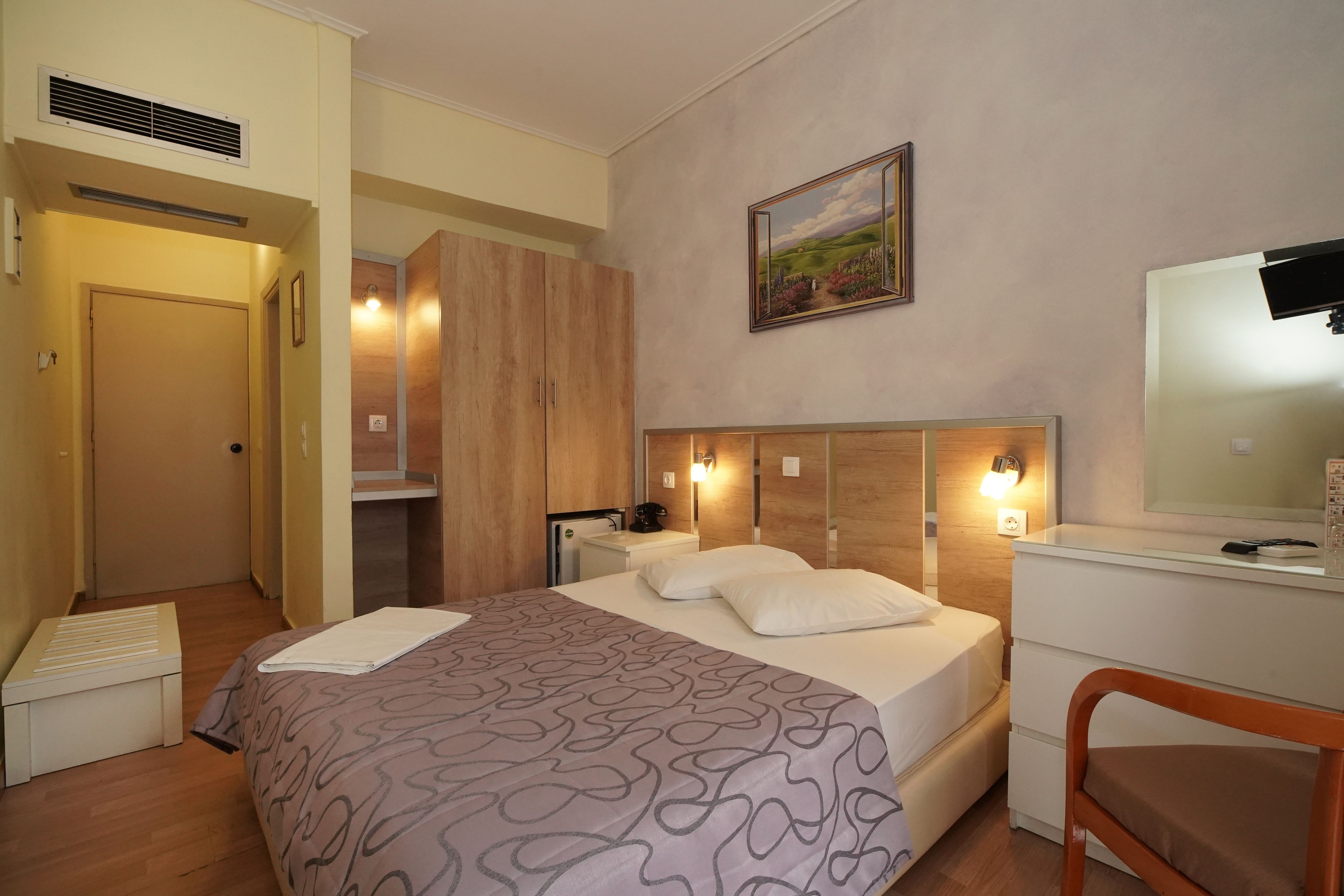 A hotel room with a bed, a desk, a chair, a wardrobe, and a painting on the wall.