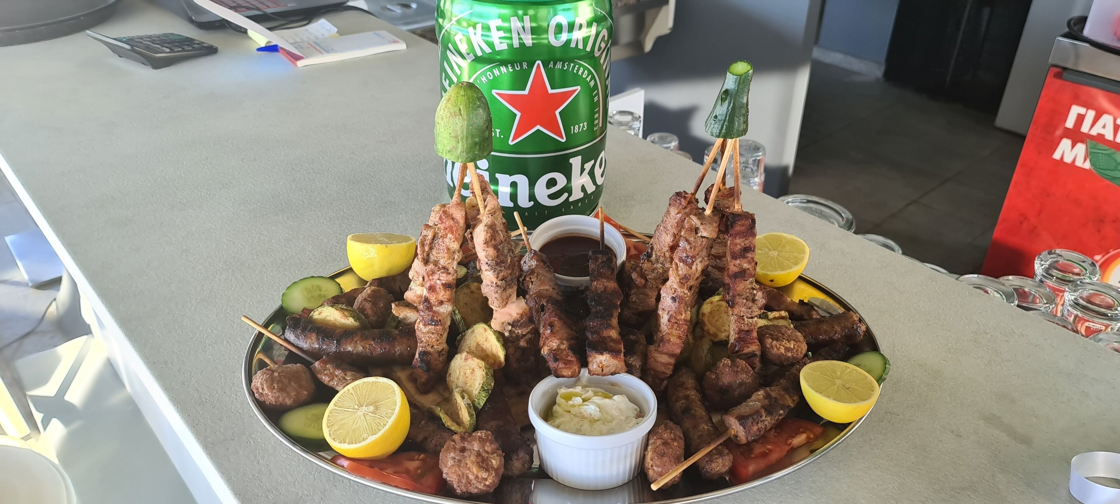 Uma mesa com uma variedade de carnes grelhadas, vegetais e uma garrafa de cerveja Heineken.