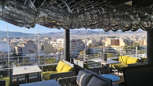 A rooftop terrace with yellow cushions, a glass balustrade, and a view of a cityscape with mountains in the background. - Ariston (Athens)
