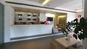 A modern reception area with a counter, shelves with plants, and a seating area with a sofa and a coffee table. - Ariston (Athens)