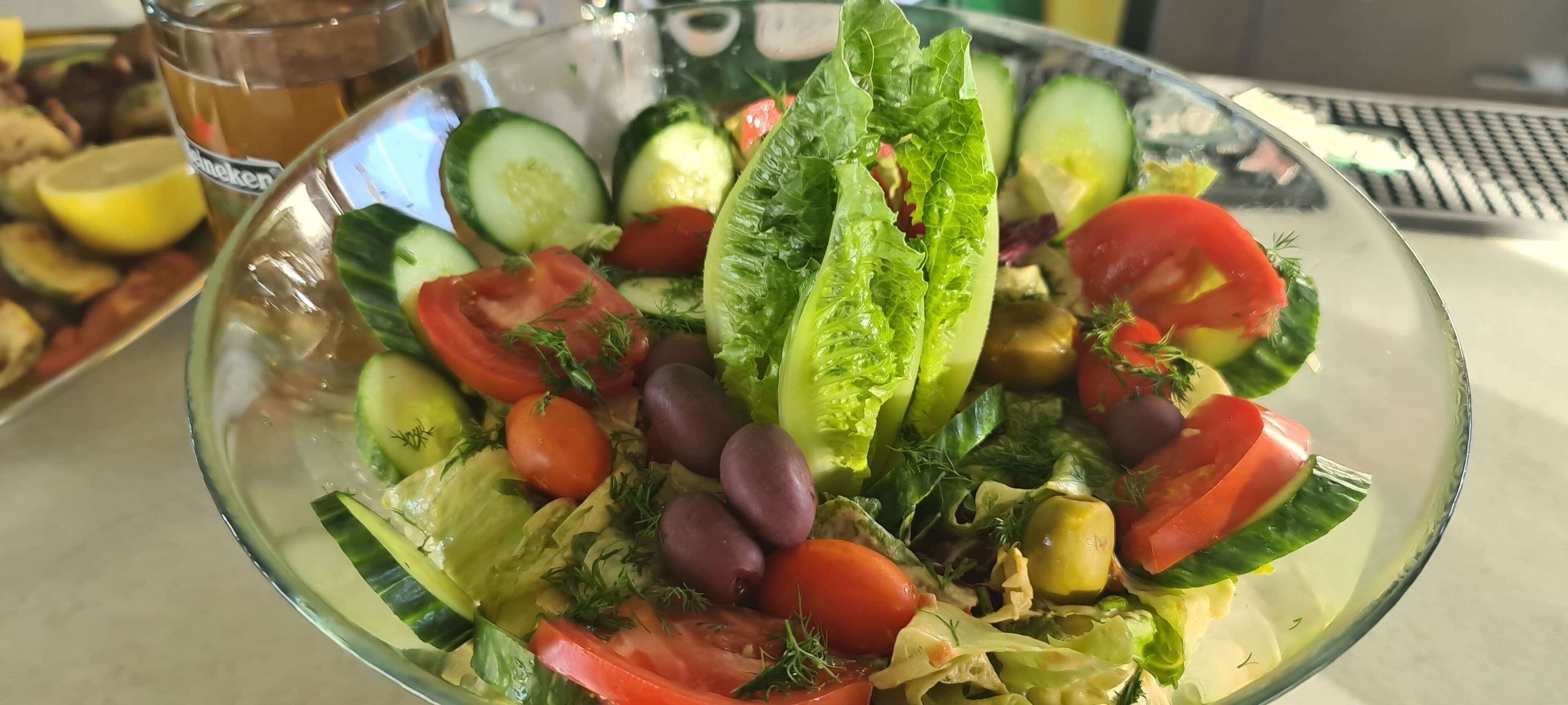 A glass bowl of salad with various vegetables and a glass of beer.