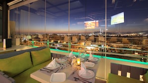 A dining area with a table set for four, a sofa, and a view of a cityscape at night. - Ariston (Athens)