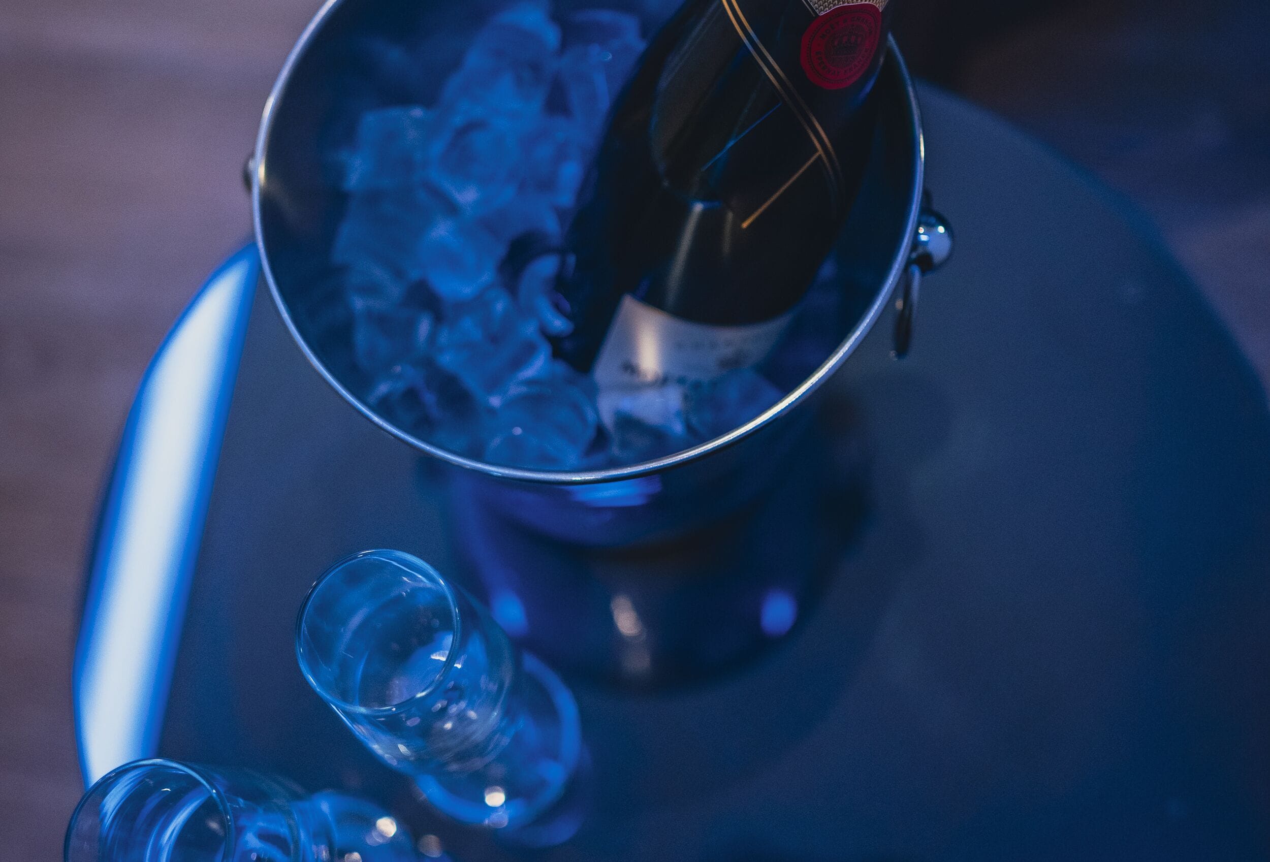 Una botella de champán en un balde de metal lleno de cubitos de hielo, con una copa y un vaso pequeño cerca.