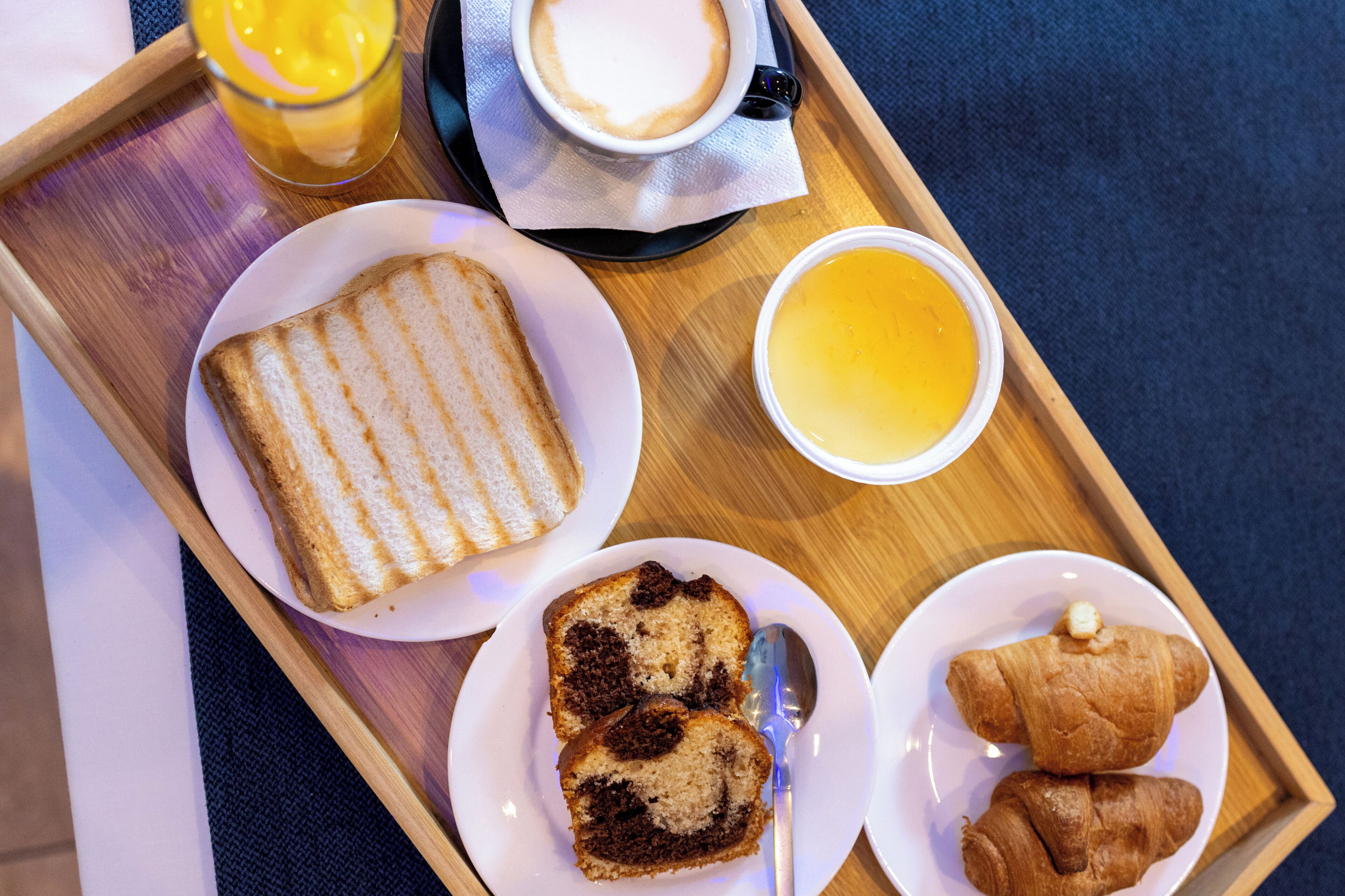 Bandeja de desayuno con tostadas, una rebanada de pastel, croissants, una taza de café y un vaso de jugo de naranja.
